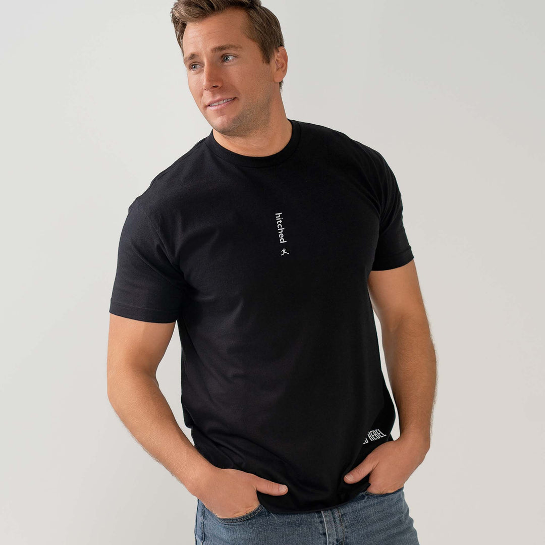 Man wearing a Bored Rebel black crewneck graphic tee and jeans. The minimalist vertical saying on his chest reads "hitched" in white ink.