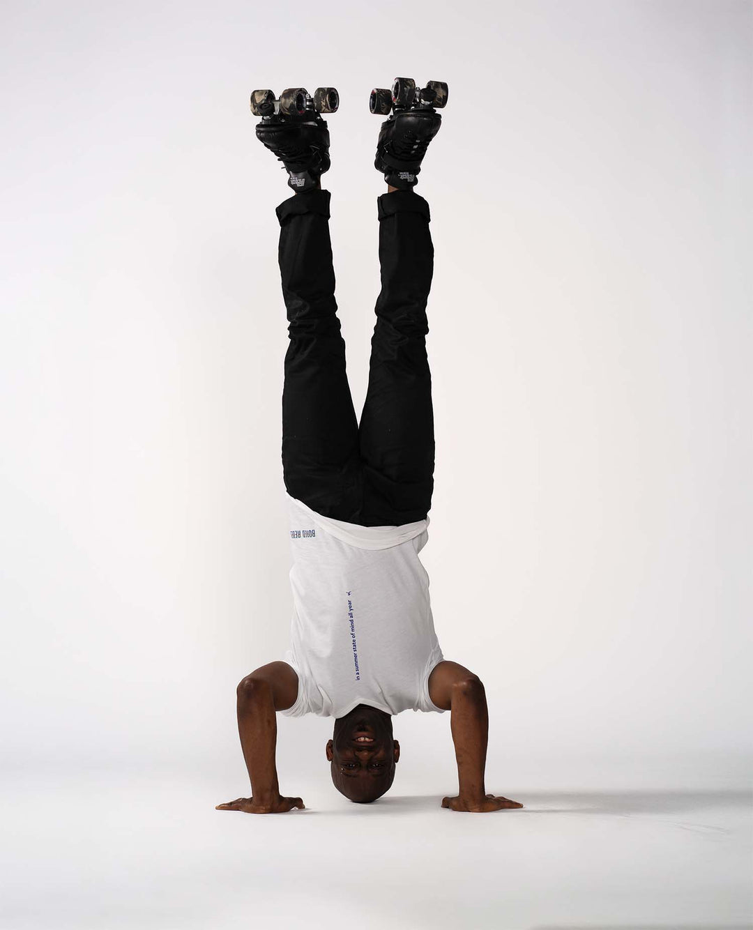 A man performing a handstand on a seamless white studio background, wearing a white Bored Rebel t-shirt, black pants, and roller skates.