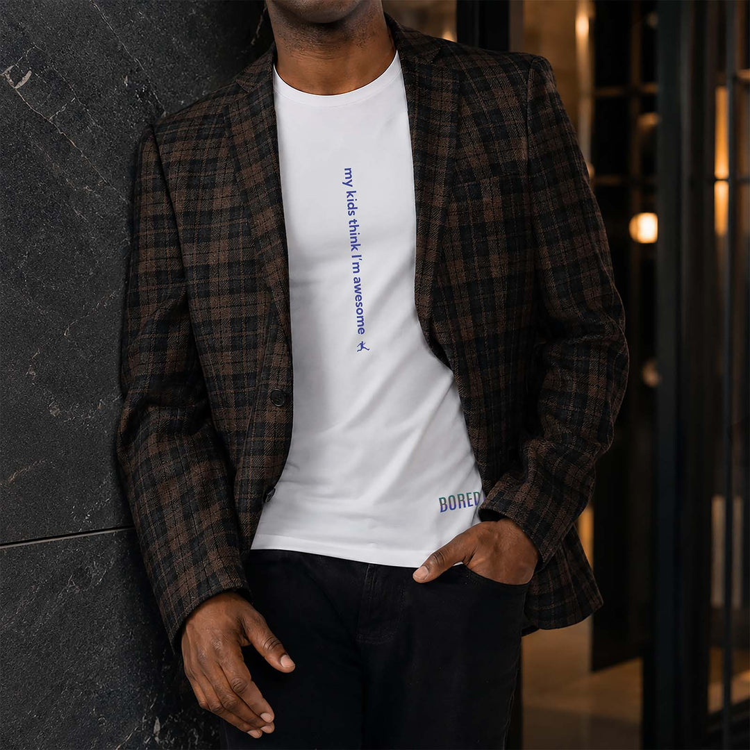A man wearing a white crewneck t-shirt with blue vertical text that reads 'my kids think I'm awesome' and a brown plaid blazer, leaning against an upscale building.