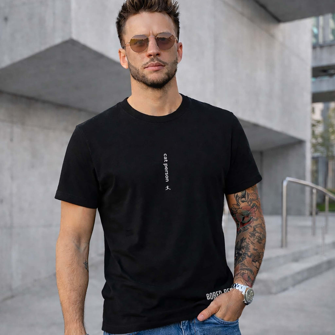Man in a black Bored Rebel minimalist graphic t-shirt and jeans standing in front of a modern concrete building. He has tattoos on one arm, a silver watch, and tinted sunglasses. His t-shirt reads 'cat person' in vertical white ink centered on his chest.