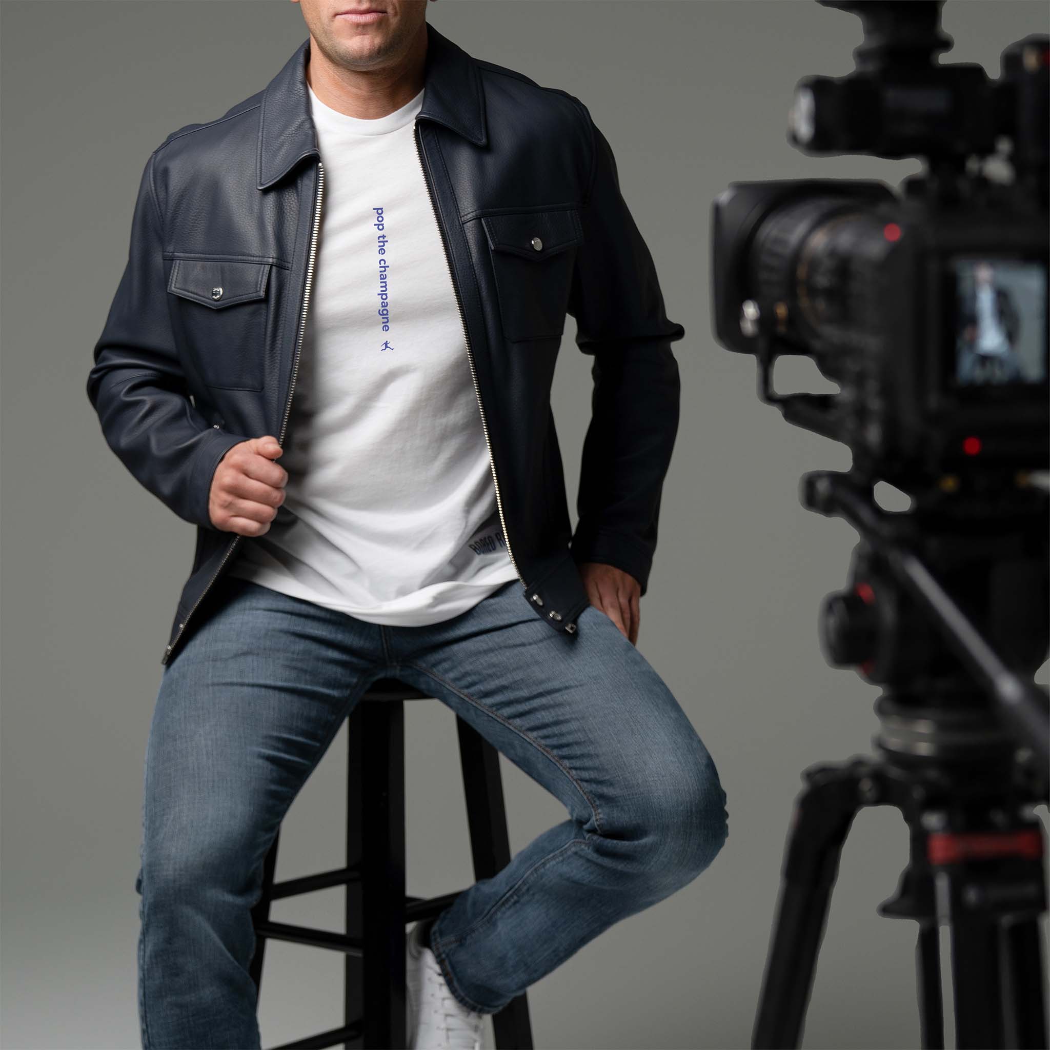 Man on a movie set is sitting on a stool wearing a navy leather jacket over a white minimalist Bored Rebel graphic t-shirt and jeans. His t-shirt reads 'pop the champagne' centered vertically on his chest. 