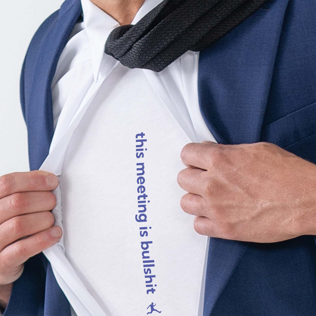 A close-up of a man opening his dress shirt, superman style, to reveal a white Bored Rebel t-shirt with blue text that reads 'this meeting is bullshit' printed vertically. 